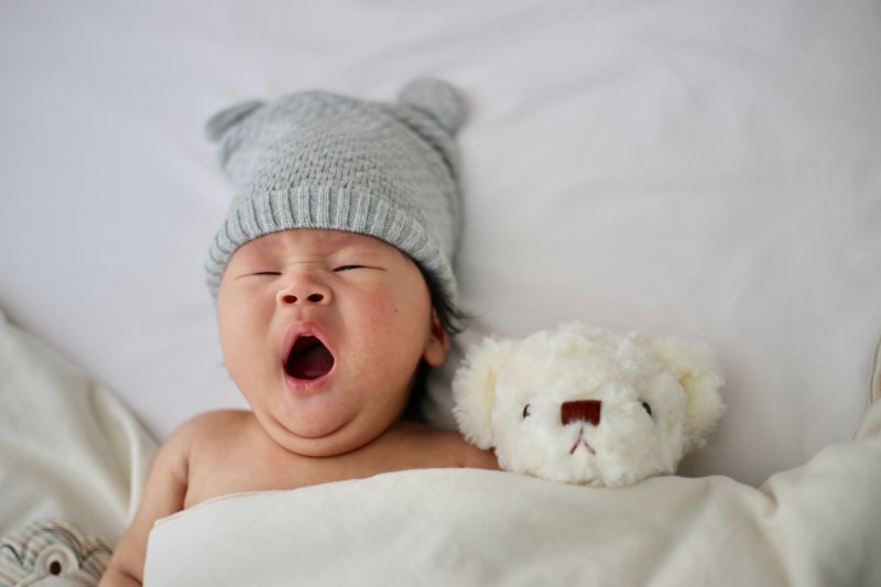 Ein gähnendes Baby mit einer grauen Strickmütze und einem weißen Teddybär