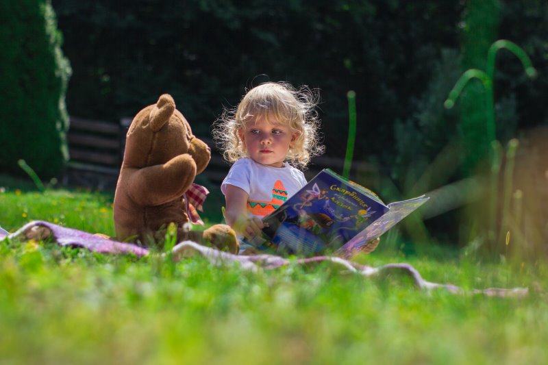 Ein blondes kleines Mädchen sitzt mit ihrem Teddybär und einem Märchenbuch auf einem geöffneten Fußsack
