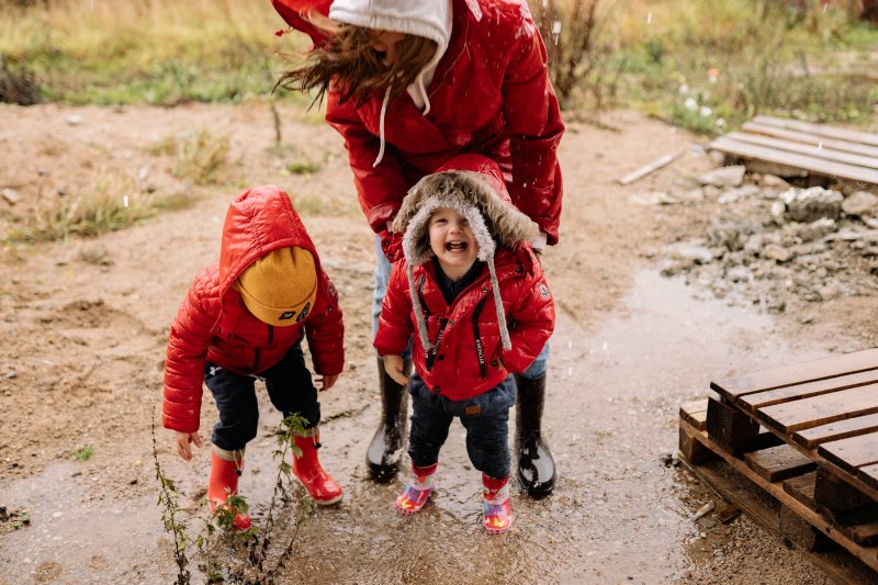 Zwei Kleinkinder mit roten Jacken und Gummistiefeln spielen mit ihrer Mutter im Matsch
