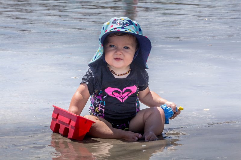 Ein Baby sitzt am Wasser im Sand mit einem Baby-Sonnenhut Ein Baby sitzt mit Sonnenhut am Strand