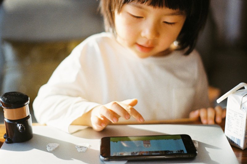 Ein kleines Mädchen sitzt an einem Tisch und spielt auf ihrem Kinderhandy mit einer App. Ein Mädchen sitzt an einem Tisch und spielt mit ihrem Kinderhandy ein Spiel