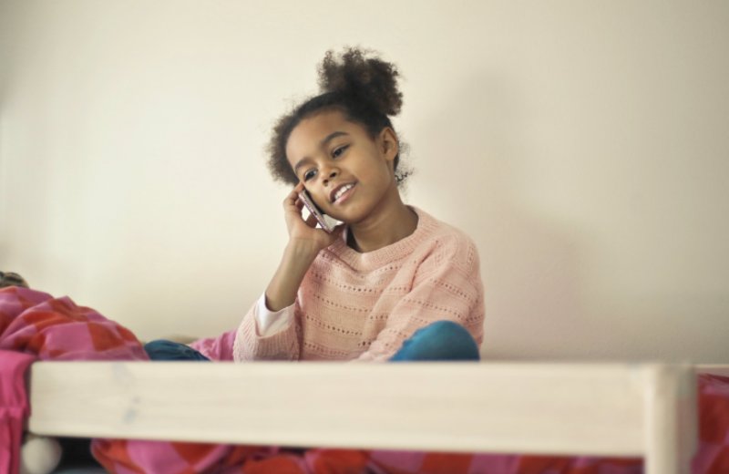 Mädchen sitzt auf einem Bett und hält sich ein Handy für Kinder gegen das Ohr. Mädchen sitzt auf einem Bett und ist am telefonieren.