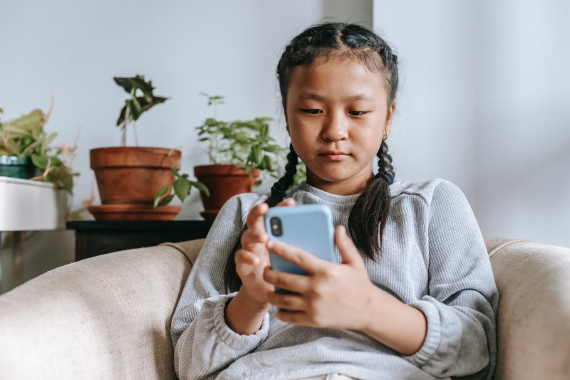 Mädchen sitzt in einem Sessel und hat ein Kinderhandy in der Hand. Ihr Blick ist konzentriert auf das Mobilgerät gerichtet. Ein junges Mädchen sitzt auf dem Sofa und hält ein Kinderhandy in der Hand.