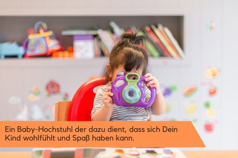 Kinder-Hochstuhl im Test Ein kleines Mädchen sitzt im Hochstuhl und hält eine lila farbige Spielzeug-Kamera vor sich.
